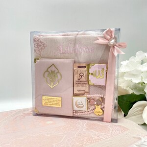 K&ouml;nnte beinhalten: Ein rosa-goldenes Geschenkset f&uuml;r ein Baby M&auml;dchen. Das Set enth&auml;lt einen Gebetsteppich, ein kleines Buch, eine Gebetsperlenkette, ein kleines Holz Ornament und eine Schachtel Tee. Der Gebetsteppich ist mit dem Namen "Adeline" bestickt.