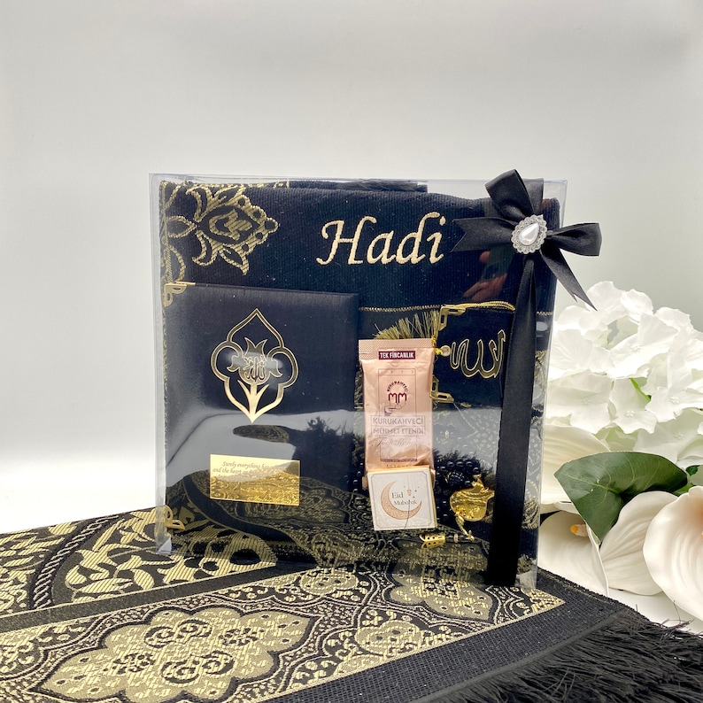May include: A black prayer rug gift set with gold embroidery. The set includes a prayer rug, a prayer mat, a small book, a bottle of perfume, and a set of prayer beads. The set is packaged in a clear plastic bag with a black ribbon and a gold-colored bow. The prayer rug has the name "Hadi" embroidered on it.
