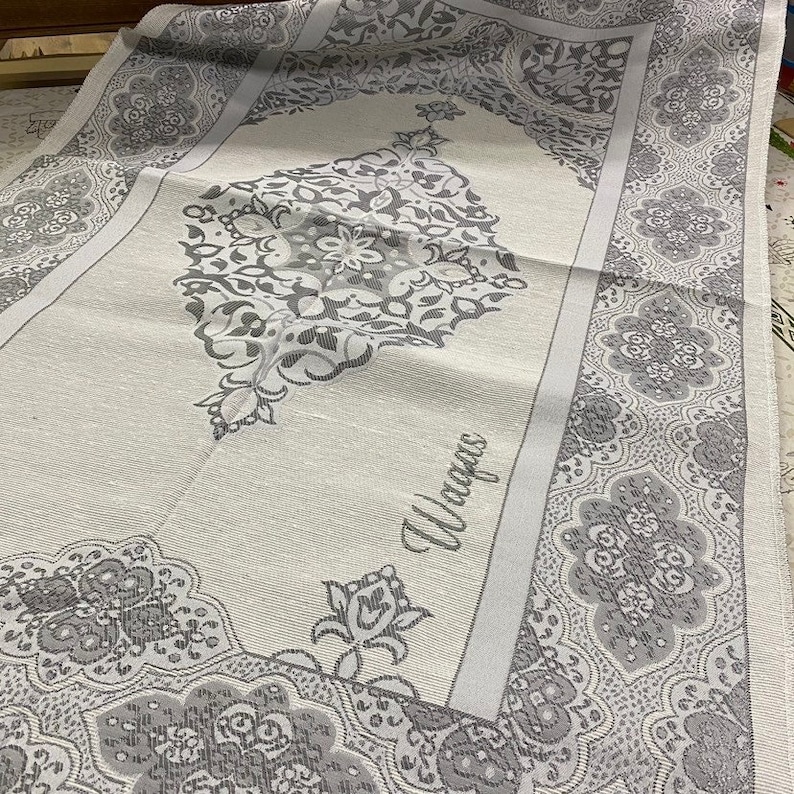 Puede incluir: Una alfombra de oraci&oacute;n blanca con un patr&oacute;n floral gris y la palabra "Wajjah" en el centro.