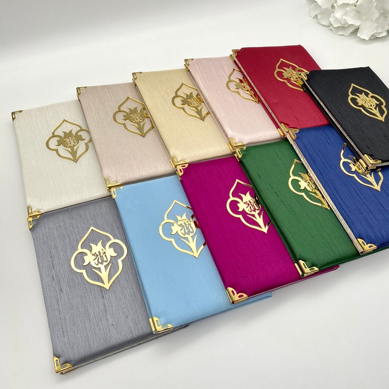 May include: A collection of 11 different colored prayer books with gold accents and a decorative design. The books are arranged in a row, with the darkest color at the back and the lightest color at the front.