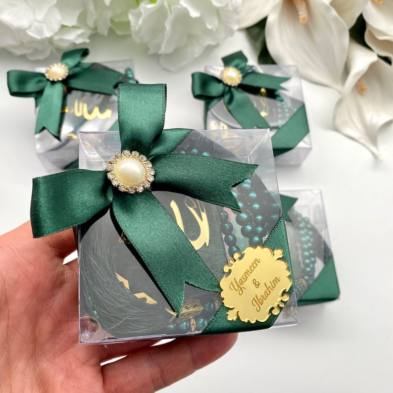 May include: Clear square boxes tied with dark green satin ribbon and pearl embellishments. Each box contains a dark green tassel, a gold-colored tag with Arabic script, and a string of dark green beads. One box has a gold tag with names.