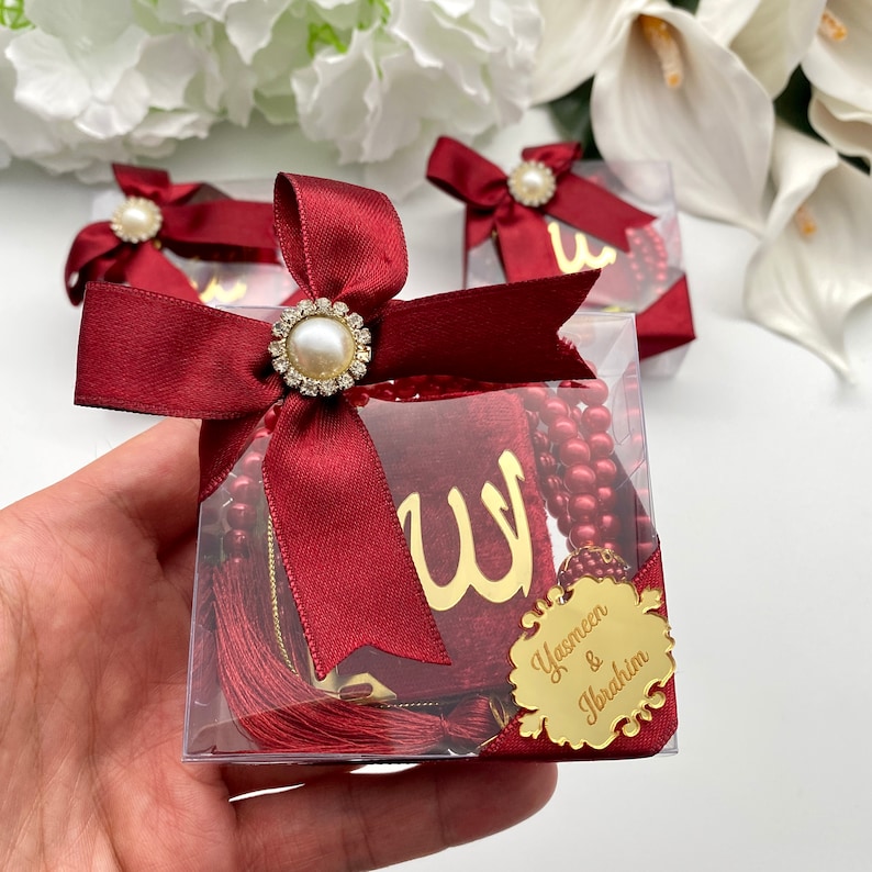 May include: Burgundy-colored gift boxes with pearl and rhinestone accents. Each box contains a prayer bead set and a small book with gold lettering. The boxes are tied with burgundy satin ribbon and a gold name tag.