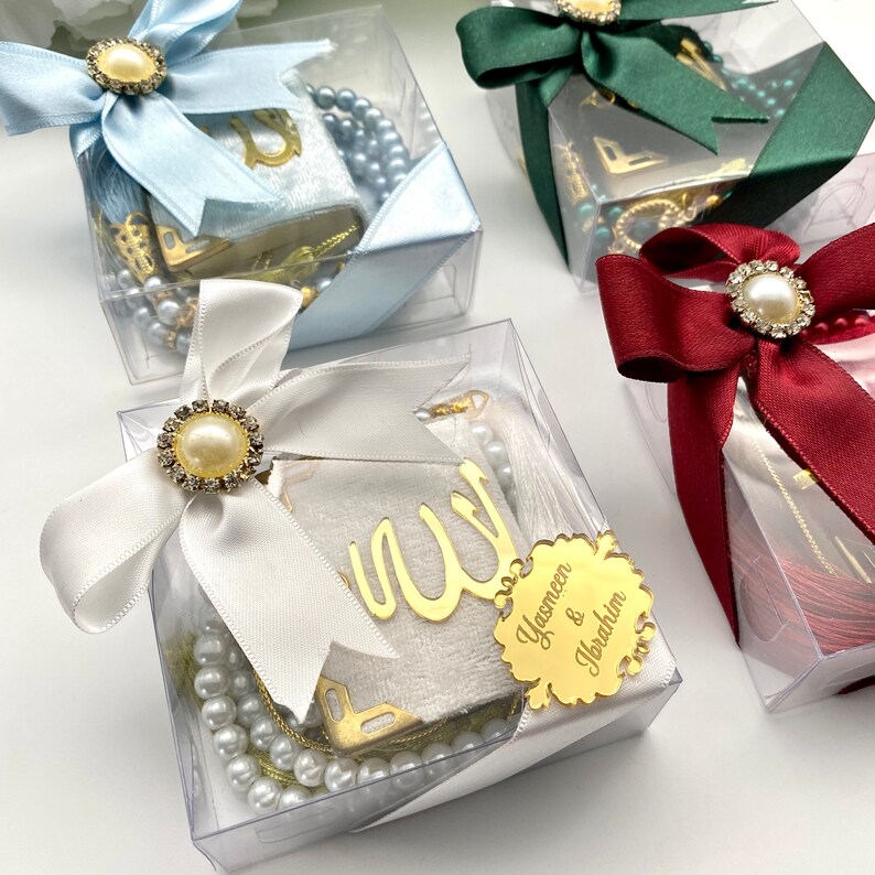 May include: Clear square boxes tied with satin ribbons in blue, white, green, and burgundy. Each box contains a small book, prayer beads, and a gold-colored charm. The ribbons are adorned with pearl and rhinestone embellishments.