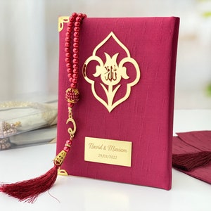 Pu&ograve; includere: Un libro di preghiere color borgogna con un fiocco dorato e un'iscrizione dorata che recita "Navid & Mariam 29/03/2022". Il libro ha un design decorativo dorato sulla copertina.