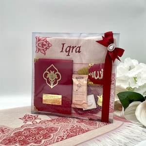May include: A gift set for a new baby girl, featuring a red and white prayer rug, a red book with gold lettering, a small red box with gold lettering, a small bag of incense, a red and gold prayer bead necklace, and a red ribbon with a gold charm. The prayer rug is personalized with the name "Iqra".