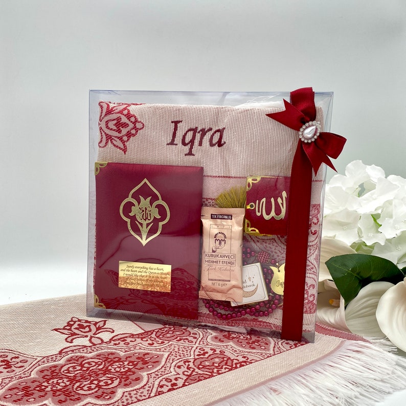 May include: A gift set for a new baby girl, featuring a red and white prayer rug, a red book with gold lettering, a small red box with gold lettering, a small bag of incense, a red and gold prayer bead necklace, and a red ribbon with a gold charm. The prayer rug is personalized with the name "Iqra".