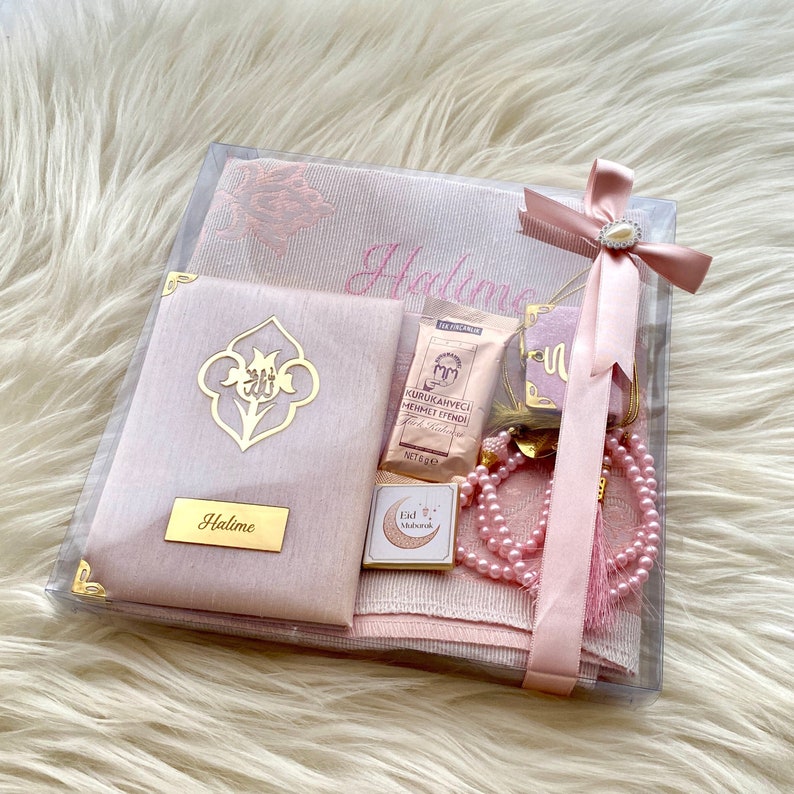 Puede incluir: Un juego de caja de regalo rosa con una alfombra de oraci&oacute;n, un libro peque&ntilde;o, un collar de cuentas, una caja peque&ntilde;a con un dise&ntilde;o de luna creciente y una peque&ntilde;a bolsa de caf&eacute;. La caja est&aacute; decorada con una cinta rosa y un adorno de perla. La alfombra de oraci&oacute;n est&aacute; bordada con el nombre "Halime".