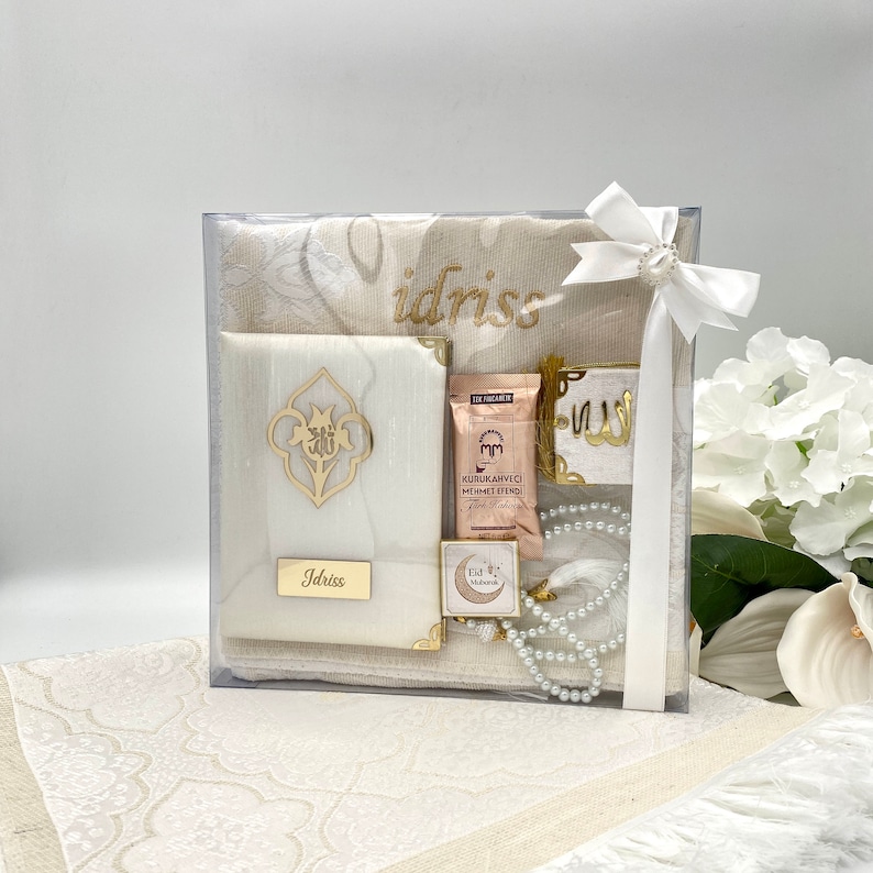 May include: A white gift box with a clear lid containing a white prayer rug, a white prayer book, a small white box with a gold crescent moon and star design, a small white box with a gold inscription, a small white box with a gold inscription, a white prayer bead necklace, and a small white box with a gold inscription. The box is tied with a white ribbon and bow. The box is sitting on a white prayer rug.