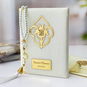 Pu&ograve; includere: Un libro di preghiere bianco con un design floreale dorato e un'iscrizione dorata che recita "Navid & Mariam 29/03/2022". Un fiocco bianco e un rosario di perle bianche sono attaccati al libro.