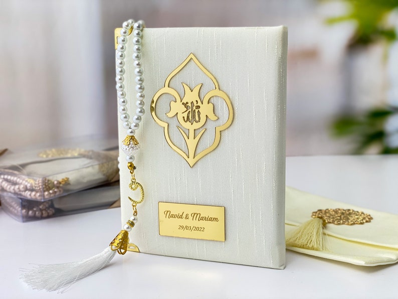 Pu&ograve; includere: Un libro di preghiere bianco con un design floreale dorato e un'iscrizione dorata che recita "Navid & Mariam 29/03/2022". Un fiocco bianco e un rosario di perle bianche sono attaccati al libro.