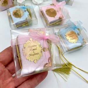 May include: A small pink velvet book with a gold inscription that reads "Bapteme Ahmed & Maghnia" in a decorative frame. The book is tied with a pink ribbon and has a gold tassel. The book is in a clear plastic box.