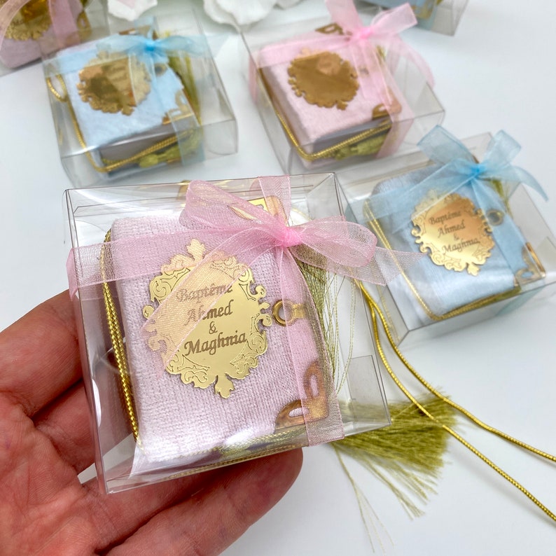 May include: A small pink velvet book with a gold inscription that reads "Bapteme Ahmed & Maghnia" in a decorative frame. The book is tied with a pink ribbon and has a gold tassel. The book is in a clear plastic box.