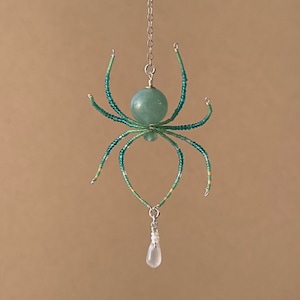 May include: A beaded spider pendant with a green and white color scheme. The spider is hanging from a silver chain and has a teardrop-shaped crystal hanging from its body.