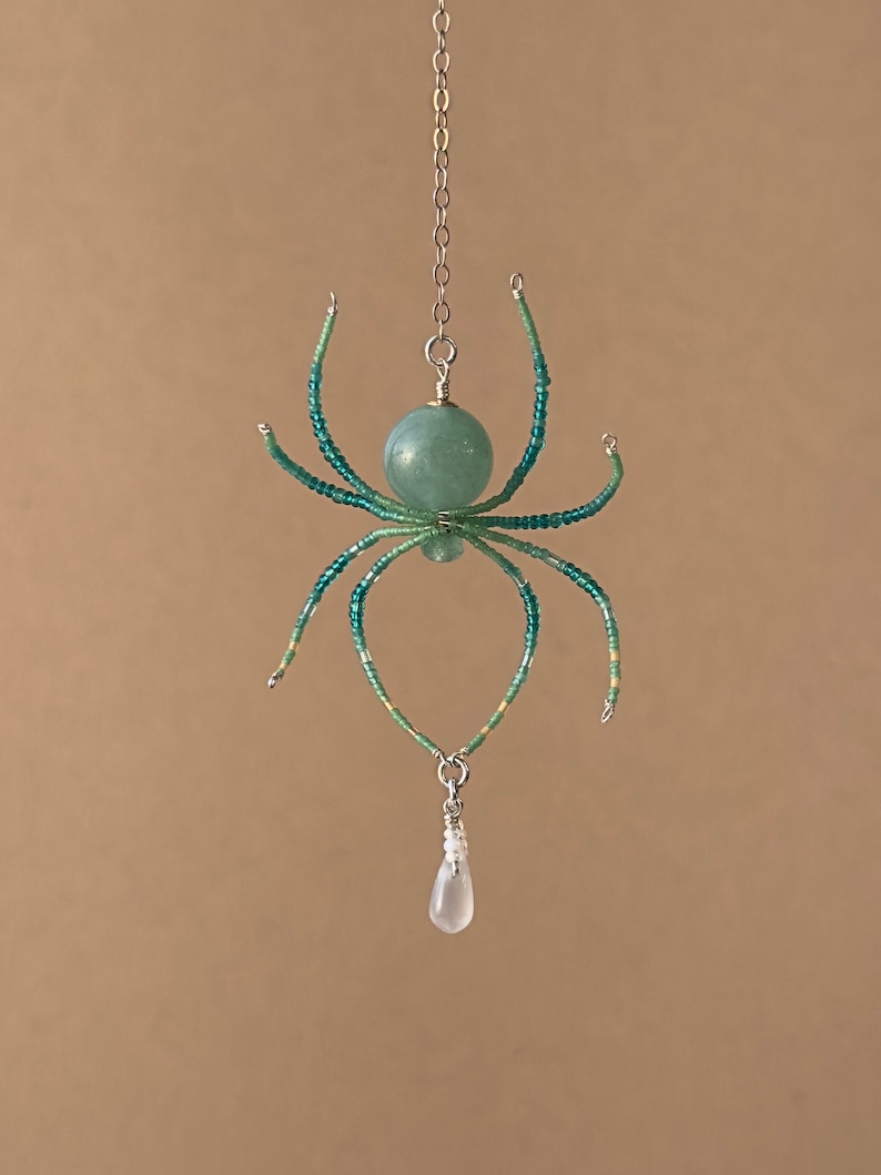 May include: A beaded spider pendant with a green and white color scheme. The spider is hanging from a silver chain and has a teardrop-shaped crystal hanging from its body.