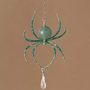 May include: A beaded spider ornament with a green and white color scheme. The spider has a round, green bead for a body and a clear crystal drop hanging from the bottom.