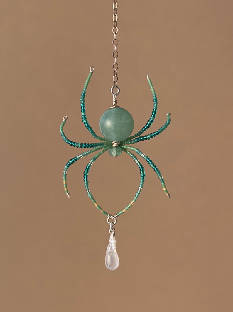 May include: A beaded spider ornament with a green and white color scheme. The spider has a round, green bead for a body and a clear crystal drop hanging from the bottom.