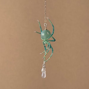 May include: A beaded spider pendant with a green gemstone body and a clear teardrop crystal hanging from the bottom. The spider is suspended from a silver chain.