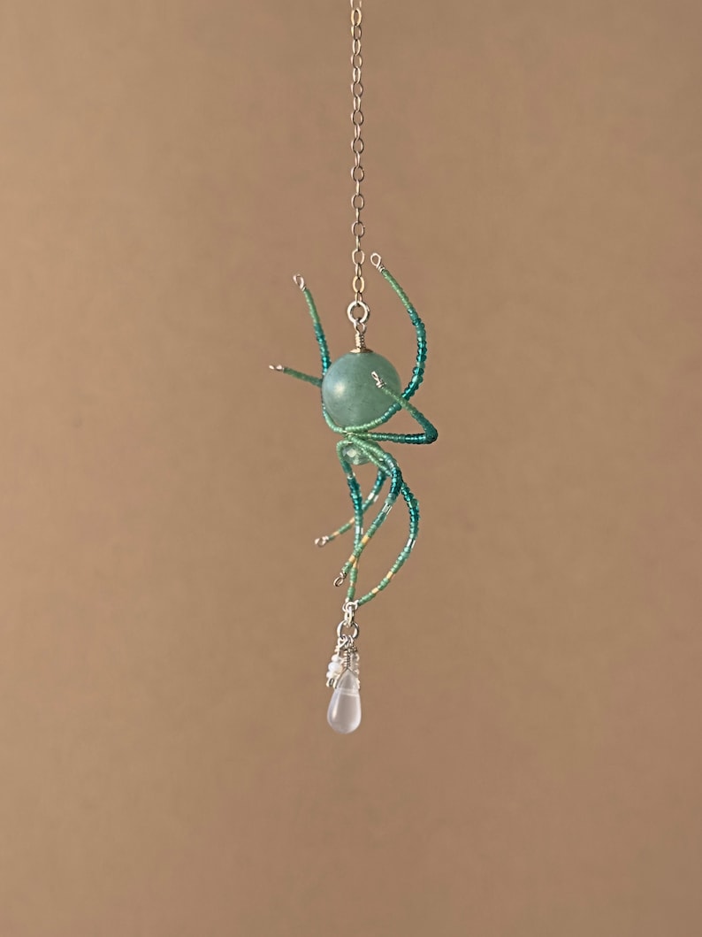 May include: A beaded spider pendant with a green gemstone body and a clear teardrop crystal hanging from the bottom. The spider is suspended from a silver chain.