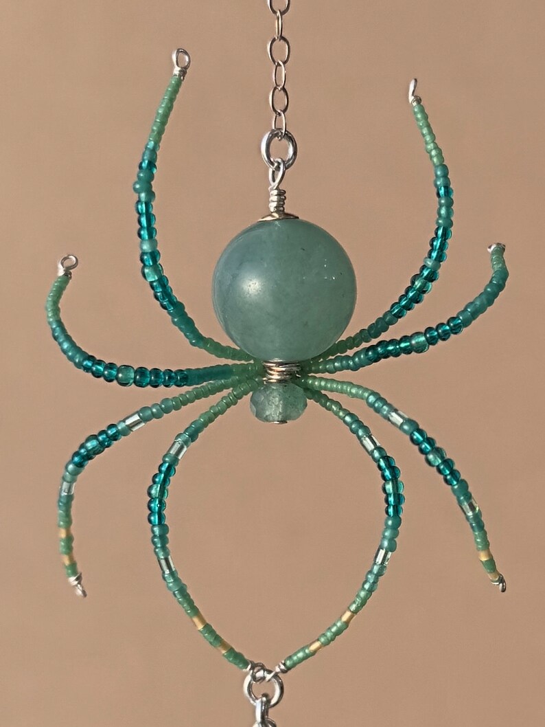 May include: A teal blue beaded spider with a green stone body hanging from a silver chain.