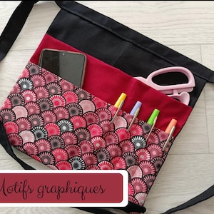 Peut inclure: Un tablier de taille noir et rouge avec un motif répétitif en forme d'éventail dans des tons de rose, rouge et noir. Le tablier comporte plusieurs poches contenant un smartphone, plusieurs stylos et des ciseaux roses. Le texte Motifs graphiques est visible.