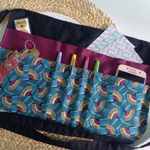 Peut inclure: Un tablier de taille noir avec une section de poche à motif d'éventails bleu sarcelle et magenta. Le tablier comprend plusieurs fentes contenant quatre stylos, un smartphone, une étiquette avec le texte Merci et un porte-clés.
