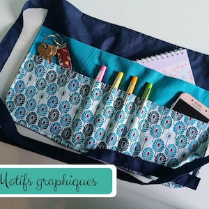 Peut inclure: Un tablier de taille bleu marine avec un panneau en tissu sarcelle orné d'un motif répétitif de plumes de paon. Le tablier comporte plusieurs poches contenant des clés, trois stylos, un carnet à spirales et un smartphone. Liens bleu marine.