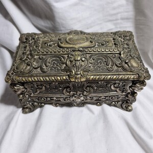 May include: Ornate silver-toned jewelry box with intricate floral and scrollwork designs. The rectangular box features a hinged lid and rests on four small feet. The box has a vintage aesthetic and is likely made of metal.