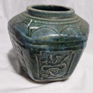 May include: A small, antique ceramic jar with a dark teal glaze. The jar has a hexagonal body with embossed floral designs and a rounded top with a decorative rim. The jar is approximately 10 cm tall.