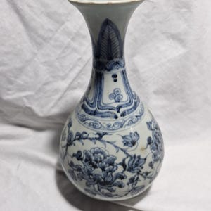 May include: A blue and white ceramic vase with a bulbous body and flared neck. The vase features intricate floral and foliate patterns in a classic blue and white colour scheme. The design includes detailed peony and leaf motifs.