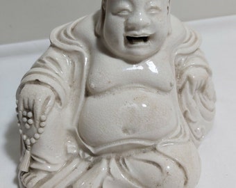 May include: White ceramic figurine of a smiling, seated Buddha. The Buddha is wearing a robe and holding a string of beads.