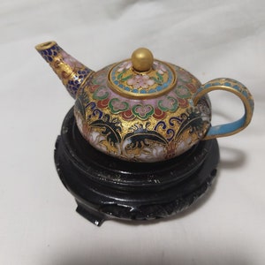 May include: A small, ornate teapot with a gold lid and colorful enamel floral designs. The teapot is sitting on a black wooden stand.