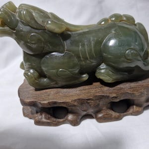 Può includere: Una scultura in giada di una creatura mitica, forse un Pixiu, in posizione reclinata. La scultura è di colore verde screziato e poggia su una base di legno marrone scuro. La creatura ha dettagli.