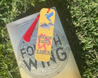 Pode incluir: Um marcador amarelo com um desenho de dragão azul e o texto "Shadow Zaddy Xaden". O marcador tem uma borla vermelha e está colocado em um livro intitulado "Fourth Wing" de Rebecca Yarros.