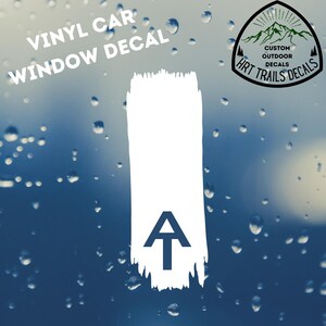 May include: White vinyl car window decal with a large white brushstroke design. The decal features a dark blue upward arrow with the letter "A" below the brushstroke. The text "VINYL CAR WINDOW DECAL" is above the brushstroke. A circular logo with mountains and the text "HRT TRAILS DECALS" is on the right.