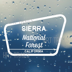 May include: A white sign with rounded corners and a blue background reads "SIERRA National Forest CALIFORNIA". The sign is on a blurry background of blue and white raindrops.