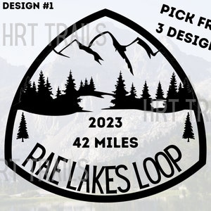 May include: Black and white illustration of a mountain range with trees and a river. The text "Rae Lakes Loop" is written in a circle around the bottom of the image. The text "2023" and "42 Miles" are written above the circle. The text "DESIGN #1" is written at the top of the image.