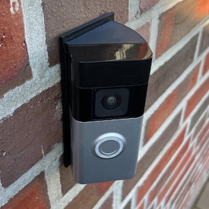 May include: A smart doorbell mounted on a red brick wall. The doorbell has a silver face with a circular button and a black camera lens. The top is black, and the device is angled slightly.