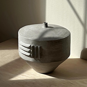 May include: A gray ceramic vase with a cylindrical body and a conical base. The vase has a small, round opening at the top and a textured, geometric design on the side. The vase is sitting on a light-colored wooden surface.