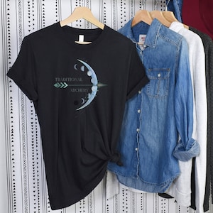 May include: Black t-shirt with a graphic of a bow and arrow, moon phases, and the words "Traditional Archery" in teal. The shirt is hanging on a wooden hanger with other clothing items, including a denim shirt and a gray sweater.