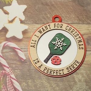May include: A round wooden ornament with a red and white border. The ornament features a green and white pickleball paddle and a red pickleball. The text on the ornament reads "All I want for Christmas is a perfect serve".