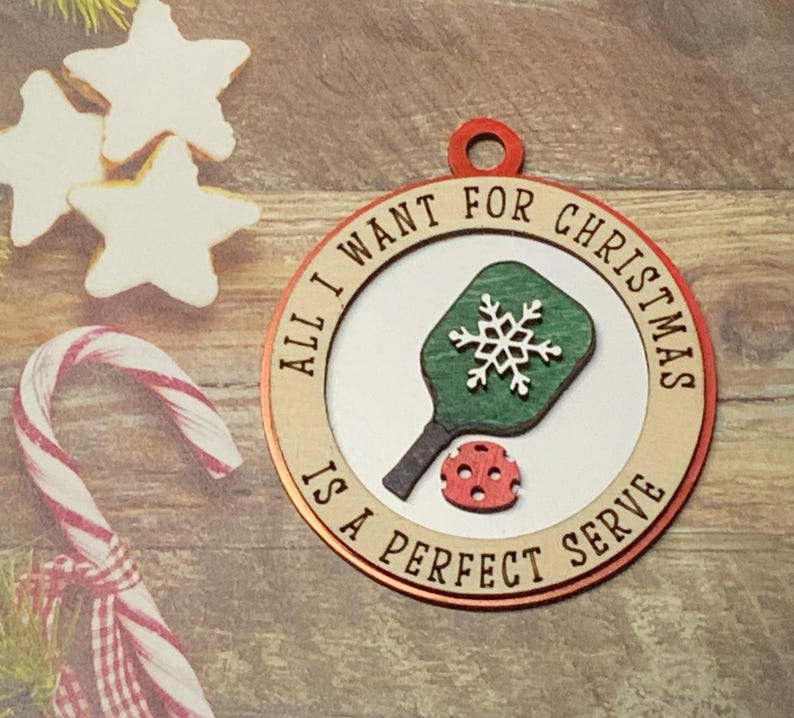 May include: A round wooden ornament with a red and white border. The ornament features a green and white pickleball paddle and a red pickleball. The text on the ornament reads "All I want for Christmas is a perfect serve".