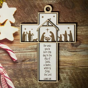 May include: Wooden cross ornament featuring a nativity scene cutout and the text "For unto you is born this day in the city of David, a Savior, which is Christ the Lord LUKE 2:11".
