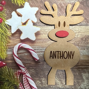 May include: A brown wooden reindeer ornament with a red nose and the name "ANTHONY" written on the body. The ornament is surrounded by Christmas decorations, including a candy cane and star-shaped cookies.