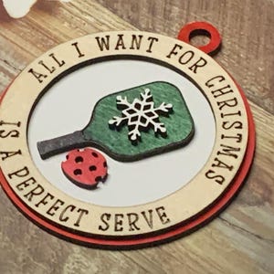 May include: A wooden Christmas ornament with a red border and the text "All I want for Christmas is a perfect serve". The ornament features a green and white pickleball paddle with a snowflake design.