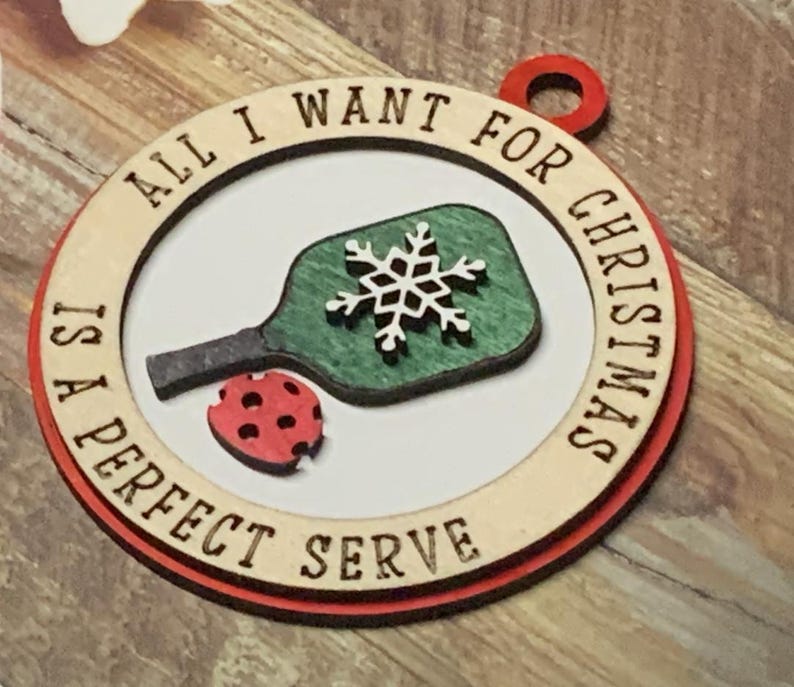 May include: A wooden Christmas ornament with a red border and the text "All I want for Christmas is a perfect serve". The ornament features a green and white pickleball paddle with a snowflake design.