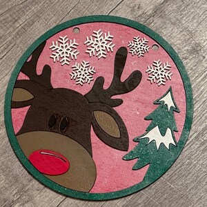 Peut inclure: Ornement de Noël rond avec un motif de renne. Le renne a des bois bruns, un nez rouge et se détache sur un fond rose avec des flocons de neige blancs et un sapin vert. L'ornement a une bordure verte.