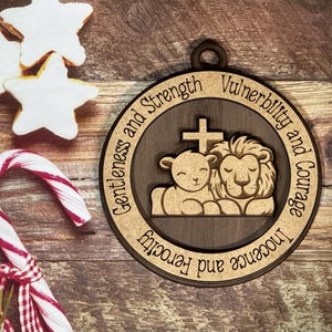 May include: A wooden ornament with a brown background and a light brown border. The ornament features a lion and a lamb with a cross above them. The text around the border reads "Innocence and Ferocity, Gentleness and Strength, Vulnerability and Courage".