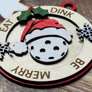 May include: A wooden Christmas ornament shaped like a pickleball with a Santa hat on top. The ornament is painted white with black holes and has the words "Eat, Dink, Be Merry" written around the edge.