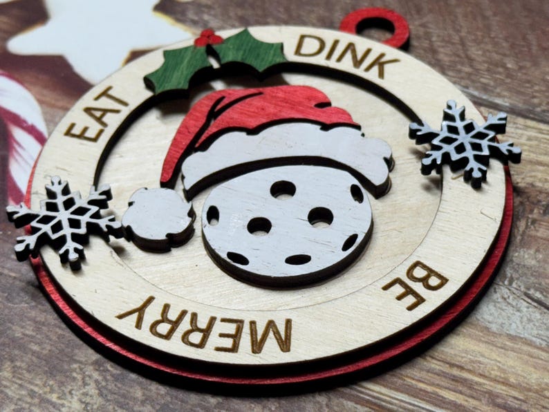 May include: A wooden Christmas ornament shaped like a pickleball with a Santa hat on top. The ornament is painted white with black holes and has the words "Eat, Dink, Be Merry" written around the edge.