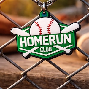 Puede incluir: Un colgante con temática de béisbol con una cadena, colgado de una valla de alambre. El colgante presenta un diseño de béisbol con costuras rojas, un fondo verde y las palabras "HOMERUN CLUB". Dos bates de béisbol blancos están cruzados detrás de la pelota.